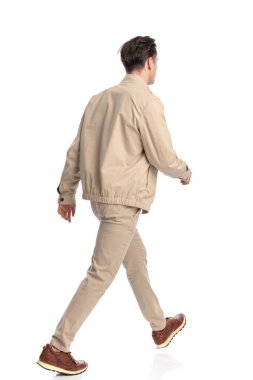 Beyaz arka planda yürüyen bej ceketli ve spor ayakkabılı sıradan bir gencin arka ve yan görünüşü.