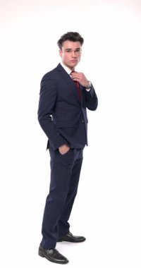 Lacivert takım elbiseli seksi bir adam kırmızı kravatı ayarlıyor ve elleri ceplerinde stüdyoda beyaz arka planda yürüyor.