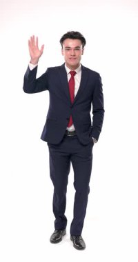 Donanma mavisi takımlı mutlu genç adamın bir elini cebinde tutarken diğeriyle el sallarken, gülümserken ve beyaz arkaplan önünde heyecanlanırken çekilmiş dikey videosu.