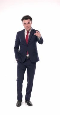 Zarif bir modacı lacivert bir takım elbise giyip beyaz arka planda dikilirken parmaklarını ileri uzatıp gülümsüyor.