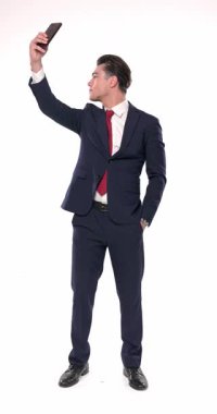 Lacivert takım elbiseli seksi, zarif bir adam telefonu kaldırıp bir elini cebinde beyaz arka planda tutarken selfie çekiyor.