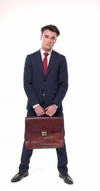 Bavulu kırmızı kravatlı lacivert takım elbiseli şık bir iş adamı beyaz arka planda dikilirken ve ciddileşirken bavulu tutuyor.