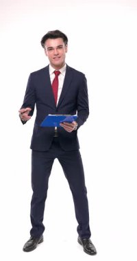 Genç işadamı, panosu ile gülümsüyor ve notlar alıyor, beyaz arka planda elini cebine koymadan önce başparmak hareketi yapıyor.