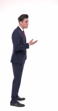 Lacivert takım elbiseli genç zarif bir adam beyaz arka planda kuyrukta beklerken önünde duran kişiyle sohbet ediyor.