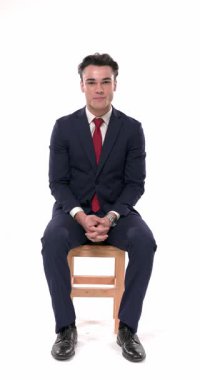 Lacivert takımlı mutlu genç adam gülümsüyor, konuşuyor ve el kol hareketleri yapıyor. Beyaz arka planda, bileği tahta koltukta otururken.