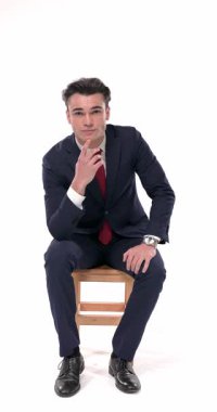 Lacivert takım elbiseli, düşünceli genç bir işadamı dizlerinin üzerinde avuç içini tutmuş, çenesine dokunuyor ve ahşap sandalyede otururken düşünüyor.