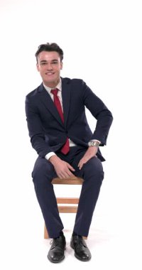 Deniz mavisi takım elbiseli mutlu bir gencin dizlerinin üzerinde dirseğini tutarken, çenesine dokunurken, gülümserken ve beyaz arka planda hayal kurarken videosu.