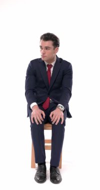 Taraf tutan lacivert takım elbiseli endişeli işadamı, beyaz arka planda otururken dizlerinin üstünde el ele tutuşup bacaklarını sallıyor.