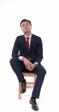 Küstah, lacivert takım elbiseli, dirseğini dizine dayamış beyaz arka planda serin bir şekilde poz verirken yan tarafa bakıyor ve çeneye dokunuyor.