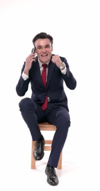 Küstah, lacivert takım elbiseli, dirseğini dizine dayamış beyaz arka planda serin bir şekilde poz verirken yan tarafa bakıyor ve çeneye dokunuyor.