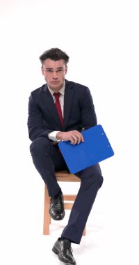 Küstah, lacivert takım elbiseli, dirseğini dizine dayamış beyaz arka planda serin bir şekilde poz verirken yan tarafa bakıyor ve çeneye dokunuyor.