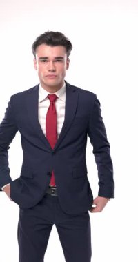 Küstah, lacivert takım elbiseli, dirseğini dizine dayamış beyaz arka planda serin bir şekilde poz verirken yan tarafa bakıyor ve çeneye dokunuyor.