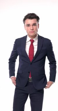 Küstah, lacivert takım elbiseli, dirseğini dizine dayamış beyaz arka planda serin bir şekilde poz verirken yan tarafa bakıyor ve çeneye dokunuyor.