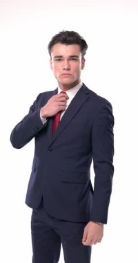 Küstah, lacivert takım elbiseli, dirseğini dizine dayamış beyaz arka planda serin bir şekilde poz verirken yan tarafa bakıyor ve çeneye dokunuyor.