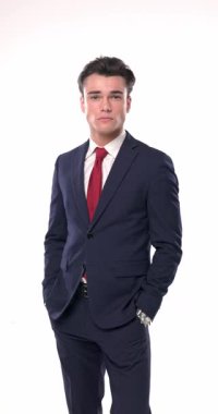 Küstah, lacivert takım elbiseli, dirseğini dizine dayamış beyaz arka planda serin bir şekilde poz verirken yan tarafa bakıyor ve çeneye dokunuyor.