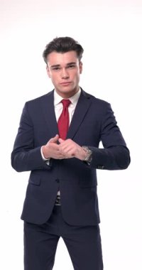 Küstah, lacivert takım elbiseli, dirseğini dizine dayamış beyaz arka planda serin bir şekilde poz verirken yan tarafa bakıyor ve çeneye dokunuyor.