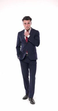 Küstah, lacivert takım elbiseli, dirseğini dizine dayamış beyaz arka planda serin bir şekilde poz verirken yan tarafa bakıyor ve çeneye dokunuyor.