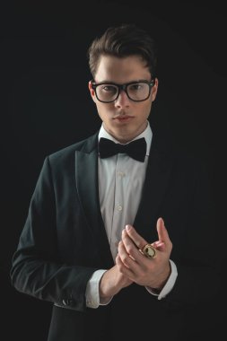 Siyah smokinli, kendine güvenen zarif bir adamın portresi. Gözlüklü, avuçlarını ovuşturan ve siyah arka planda poz veren.