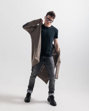 Gözlüklü havalı adam gri arka planda kapüşonluyu ayarlarken kendine güvenen bir şekilde poz veriyor.