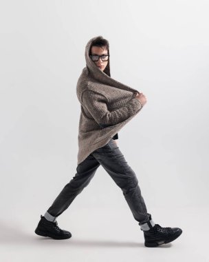 Gözlüklü, örgü kapüşonlu ve gri arka planda yürüyen havalı moda adamı.
