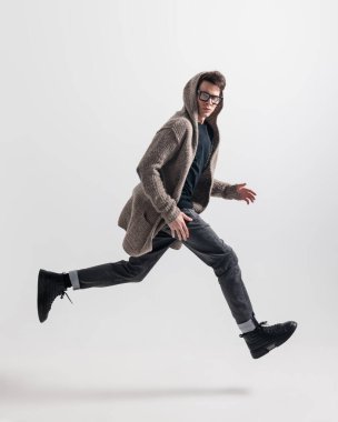 Gözlüklü, örgülü kapüşonlu ve gri arka planda havaya zıplayan dinamik modacının yan görünüşü