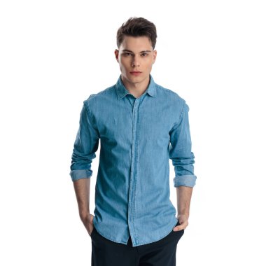 Önüne bakıp beyaz arkaplan önünde poz verirken ceplerinde el ele tutuşan kendinden emin genç bir adam.