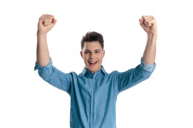 Kot pantolonlu heyecanlı genç adam kafasının üstünde yumruklar tutuyor ve beyaz arka planda gülüyor.