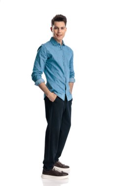 Mavi kot gömlekli yakışıklı genç adam ceplerinde el ele tutuşuyor ve beyaz arka planda dururken gülümsüyor.