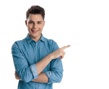 Mavi gömlekli mutlu bir gencin portresi. Beyaz arka planda dikilirken parmaklarını sağa sola gösteriyor ve gülümsüyor.