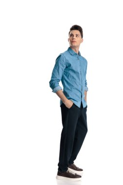 Mavi kotlu gömlekli yakışıklı genç adam elini cebine sokup beyaz arka planda dikilirken yukarı bakıyor.