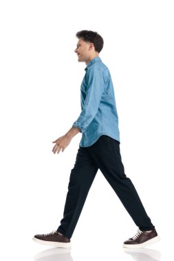 Mavi kot gömlekli mutlu genç adam beyaz arka planda yürüyor ve gülümsüyor.