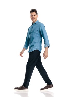 Mavi kot gömlekli sıradan bir genç adam mutlu ve gülümsüyor, beyaz arka planda yürüyor.