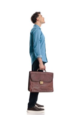 Mavi kot pantolonlu, bavulu tutan, ayakta duran ve beyaz arka plana bakan meraklı genç adamın yan görüntüsü.