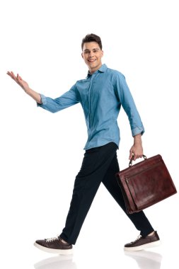 Mavi kot gömlekli mutlu genç adam beyaz arka planda yürürken bavulunu yana doğru sunuyor.