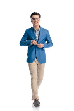 Mavi ceketi ilikleyen ve beyaz arka planda yürürken gülümseyen gözlüklü yakışıklı genç adam.