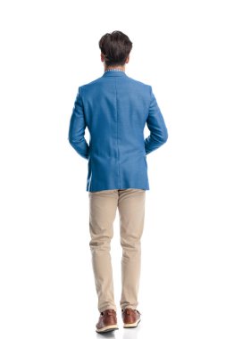 Mavi ceketli, beyaz arka planda yürüyen, Chino pantolonlu ve spor ayakkabılı genç adam manzarasının arkasında.