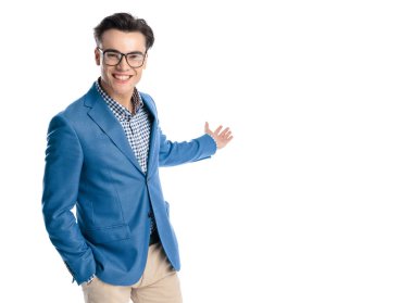Mavi ceketli gururlu genç adam, beyaz arka planda gülümserken elini yana doğru gösteriyor.