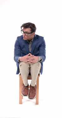 Her iki tarafa bakıp beyaz arka planda oturan gözlüklü, hareketli genç adamın dikey videosu.