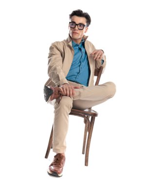 Kendine güveni olan, gözlüklü, tahta koltukta oturan, bacağı dizinde ve beyaz arka planın önünde duran bir adam.