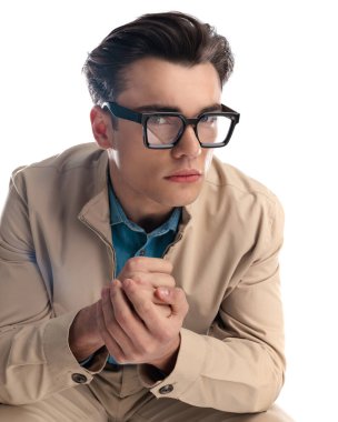 Siyah gözlüklü, kendinden emin genç bir adam. Önüne bakıp beyaz arka planda otururken yumruklarını ovalıyor.