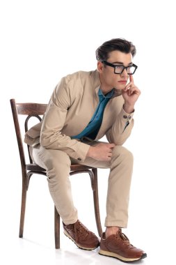 Gözlüklü düşünceli genç adam, beyaz arka planda otururken çenesine dokunup düşünüyor.