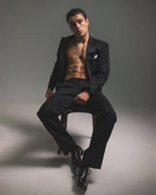 Siyah smokinli seksi genç iş adamı gri arka planda havalı bir şekilde poz verirken sandalyeye oturuyor ve bakıyor.