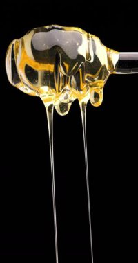 Bal kaşığının üzerine bal dökülüyor ve ipeksi dokusunu, doğal ışığını ve zengin altın rengini vurguluyor.