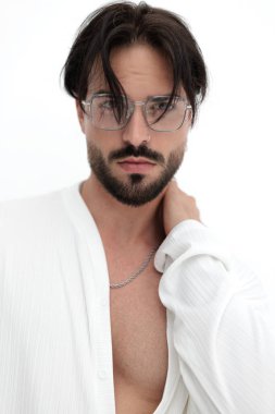 Beyaz gömlekli, gözlüklü, önüne bakan, el ele tutuşan ve beyaz arka planda poz veren kendinden emin bir adam.