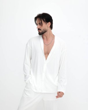 Beyaz kostümlü çekici sakallı bir adam beyaz arka planda elleri ceplerinde poz veriyor.