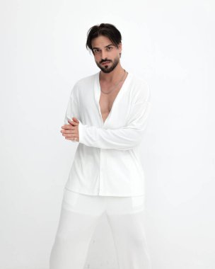 Beyaz kostümlü yakışıklı genç adam avuçlarını ovuyor ve beyaz arka planda dikilirken ileriye bakıyor.