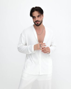 Sakallı genç bir adam beyaz arka planda ilerlerken yüzükler takıyor ve beyaz kostümlü poz veriyor.