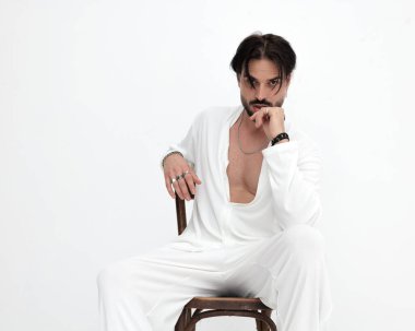 Beyaz kostümlü seksi manken portresi beyaz arka planda tahta koltukta otururken dudaklara dokunuyor.