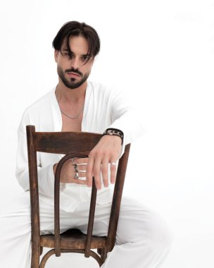 Beyaz kostümlü havalı bir adamın portresi. Dirseğini sandalyeye koyup beyaz arka planda oturuyor.