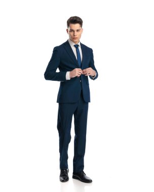 Zarif genç işadamı lacivert takımını ilikliyor ve beyaz arka planın önünde yürüyor.